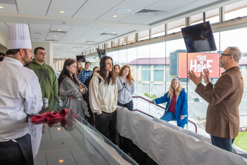 A group of high school students stand in a room overlooking the Virginia Tech football field listening to a man in a suit talk.