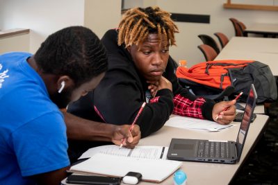Two male students sit working on physics worksheets as they check their work on a laptop.