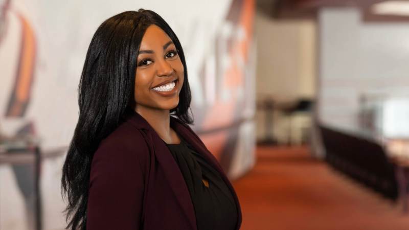 Ashley Offutt stands at an angle to the camera, smiling with her long hair behind her shoulders. The background, which appearance to be a conference or event center, is blurred.