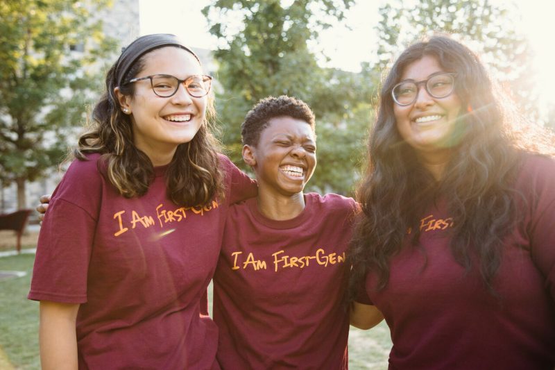 Three students smile, nearly laughing, as they stand together wearing matching maroon shirts with orange script reading "I am first-gen." Leafy trees stand in the background.