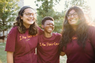 Three students smile, nearly laughing, as they stand together wearing matching maroon shirts with orange script reading "I am first-gen." Leafy trees stand in the background.