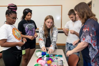 A group of teens and a teacher gather around a desk holding spools of colorful yarn.
