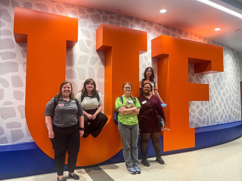 A small group of high schoolers sit on giant orange letters U.F.